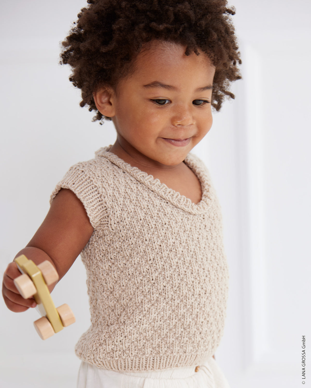 Barn i strikket slipover med V-hals, strukturmønster og ribkanter i Cool Wool Big fra Lana Grossa i farven Beige (945).