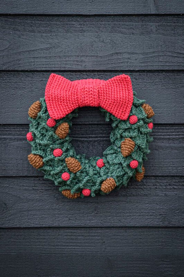 Hæklet julekrans med grønne blade i krokodilleskæl, stor rød sløjfe, røde bær og brune kogler i Cotton 8/4 fra Mayflower.